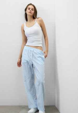 Ung kvinde iført hvid tanktop og lyseblå, stribede pyjamasbukser står op ad en hvid hjørnevæg.