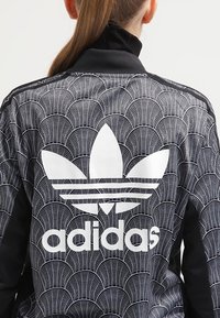 Černá sportovní bunda s texturovaným vzorem překrývajících se oblouků. Bílé logo Adidas je výrazně umístěno na zadní straně.