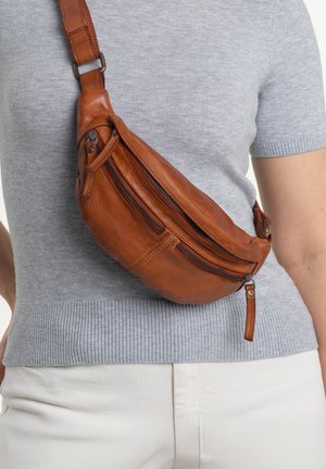 Brown leather sling bag with two zippered compartments, curved shape, and adjustable strap, worn over a light gray shirt. Textured surface.