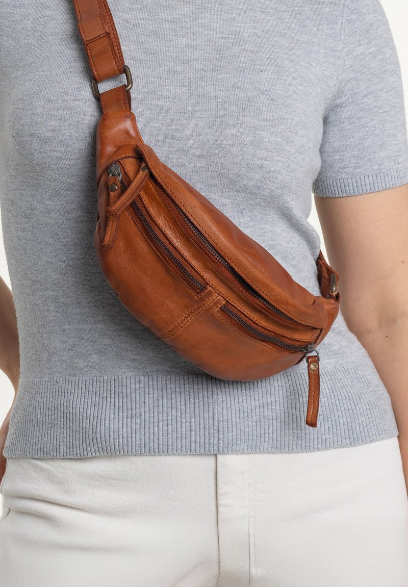 Brown leather sling bag with two zippered compartments, curved shape, and adjustable strap, worn over a light gray shirt. Textured surface.