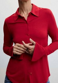 Chemise rouge à manches longues en tissu doux. Elle possède un col classique, une fermeture à boutons sur le devant, et des poignets évasés. Ourlet légèrement arrondi.