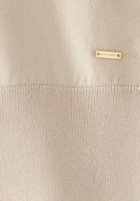 Maglione lavorato a maglia beige con una texture liscia e a coste, dotato di un'etichetta in metallo dorato con la scritta "HOLLY & WHITE" sul davanti.