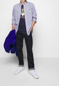 Mies, joka on pukeutunut raidalliseen siniseen paitaan, jossa on tummansininen karhukuva t-paitana, tummiin farkkuihin, jotka ovat käännetty nilkoista, ja valkoisiin lenkkareihin, pitää kädessään purppura takkia.