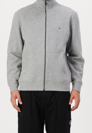 Man wearing a light grey zip-up sweatshirt with side pockets and black pants, shown from neck to hips against a white background.