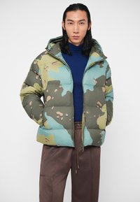 Mees pika musta juustega, kelles on seljas sinine kõrge kaelusega särk, roheline ja sinine kamuflaažiga sulejope ning pruunid siduripüksid valgel taustal.