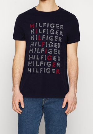 Mand iført en sort t-shirt med gentaget "HILFIGER" tekst; lodrette bogstaver i rødt staver "HILFIGER" ned ad fronten.