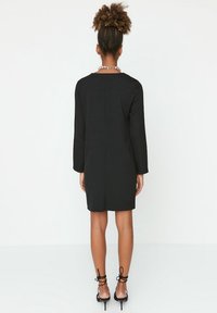 Kvinna med lockigt hår i en hög knut, klädd i en svart långärmad klänning, svarta remsandaler och ett pärlhalsband, vänd bort från kameran.