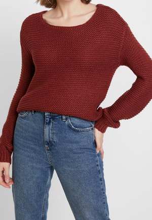 Femme portant un pull en maille bordeaux texturé à manches longues et un jean bleu taille haute, main posée sur la hanche sur un fond uni.
