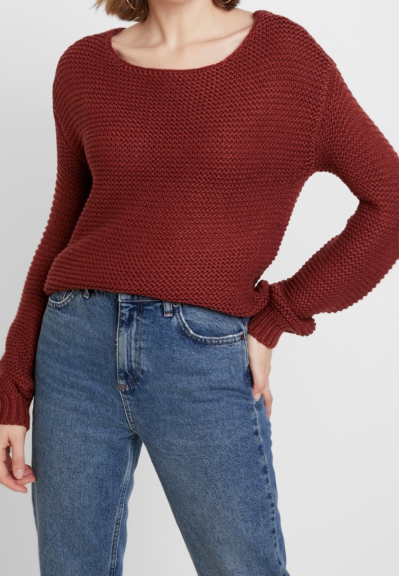 Femme portant un pull en maille bordeaux texturé à manches longues et un jean bleu taille haute, main posée sur la hanche sur un fond uni.