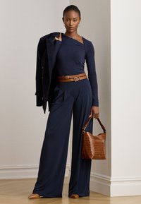 Moteris vilki tamsiai mėlynas plačias kelnes ir ilgom rankovėm marškinėlį su dviguba ruda diržu, laiko rudą odinę rankinę ir tamsiai mėlyną švarką per petį.