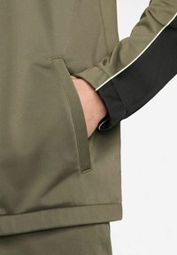 Chaqueta verde y negra con un bolsillo con cremallera y detalles en las mangas en contraste. La tela es suave con un acabado ligeramente brillante.