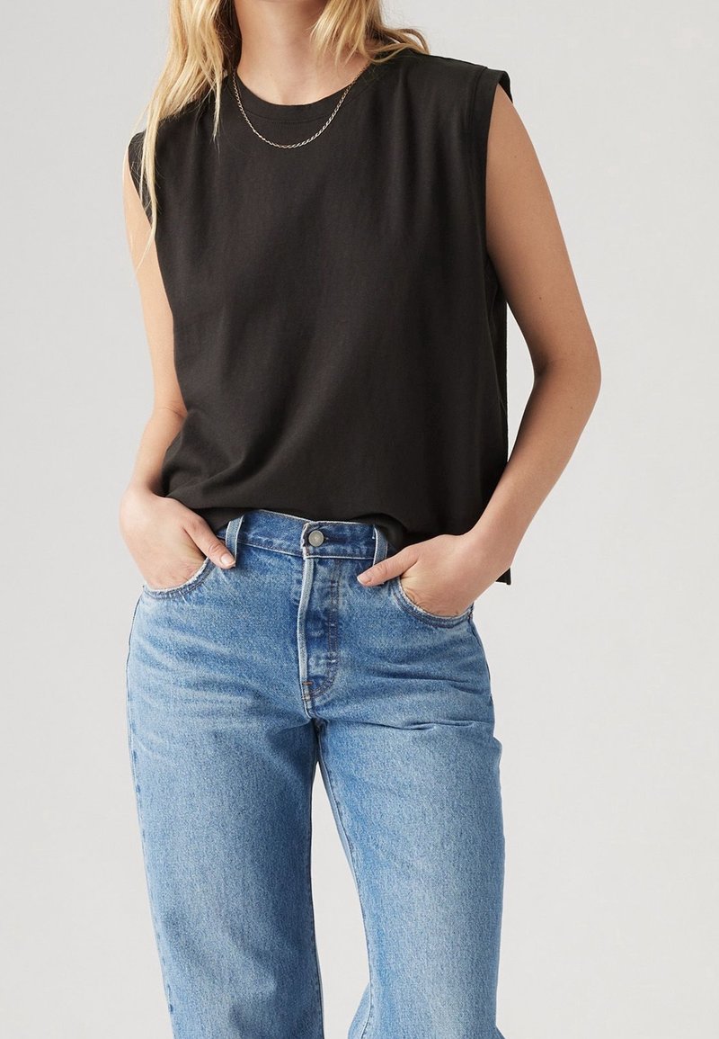 Personne portant un haut noir sans manches, un jean bleu clair taille haute et un fin collier chaîne, mains dans les poches, face à l'avant.