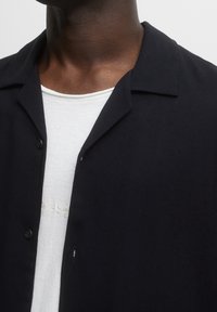 Camicia nera a maniche corte con bottoni in un tessuto leggero, dotata di colletto e abbottonatura, indossata sopra una maglietta bianca con ricami sottili.