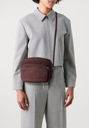 Borsa a tracolla in suede marrone con due scomparti con zip e una lunga tracolla, abbinata a una camicia a quadri e pantaloni coordinati.