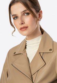 Beige Lederjacke mit einem Reverskragen, diagonaler Reißverschlussöffnung und dezenten Stickdetails, getragen über einem cremefarbenen Rollkragenpulli.
