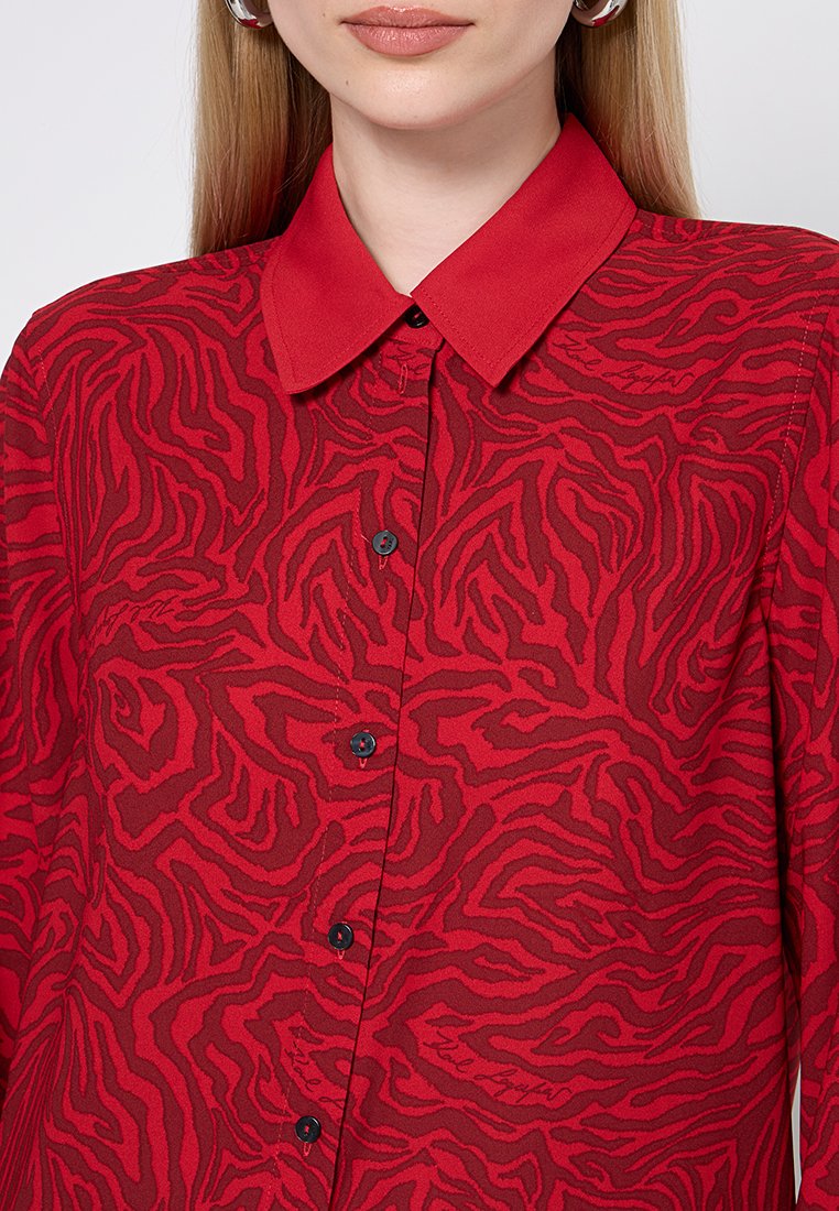 Chemise rouge avec un motif zèbre tigre, fermeture à boutons sur le devant et col. Le tissu a une texture lisse avec un subtil éclat. Boutons noirs en accent.