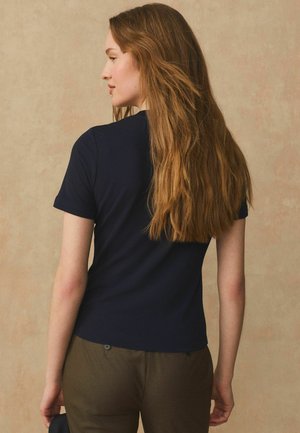 Hosszú világosbarna hajú nő, sötétkék rövid ujjú ingben és olívazöld nadrágban, balra nézve, bézs háttér előtt.