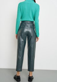 Žena oblečená v tyrkysovém žebrovaném svetru zasunutém do vysokých tmavě zelených kožených kalhot s černými platformovými botami, stojící čelem ke bílé zdi.