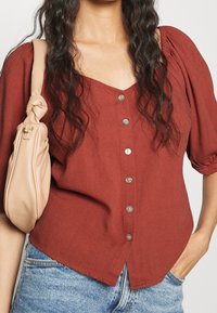 Rostfarbige Bluse mit V-Ausschnitt, kurzen Puffärmeln und sechs silbernen Knöpfen auf der Vorderseite; kombiniert mit einer hellbeigen Handtasche.