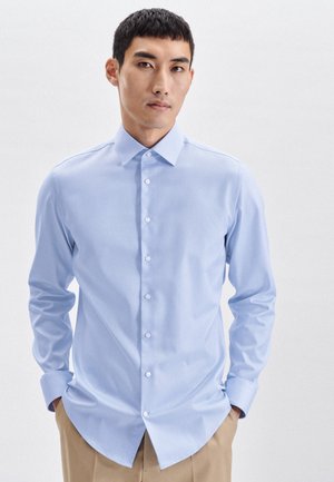 Helleblaue Langarmbluse mit Button-Down-Kragen, glatter Textur, weißen Knöpfen und abgerundetem Saum, kombiniert mit beigen Hosen.
