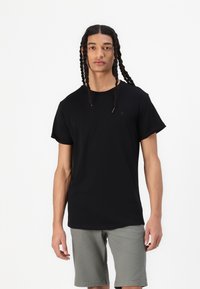 Tricou negru din bumbac cu guler rotund și mâneci scurte, având un logo discret pe piept. Purtați cu pantaloni scurți gri.