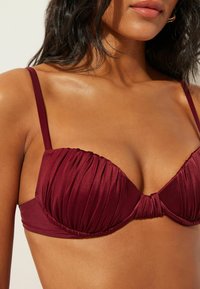 Bordowy top bikini z plisowanym wzorem, lekko wyściełanymi miseczkami i cienkimi regulowanymi ramiączkami. Z centralnym węzłem jako akcentem. Gładka tekstura.