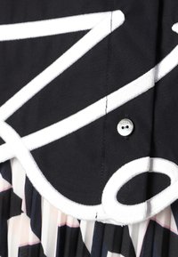 Tessuto nero con design grafico bianco e dettagli plissettati rosa. Presenta un bottone argentato per la chiusura. Morbido al tatto.