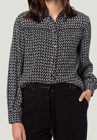 Chemise noire et blanche à boutons avec un motif géométrique, manches longues, et une poche poitrine, associée à un pantalon noir avec un cordon de serrage.