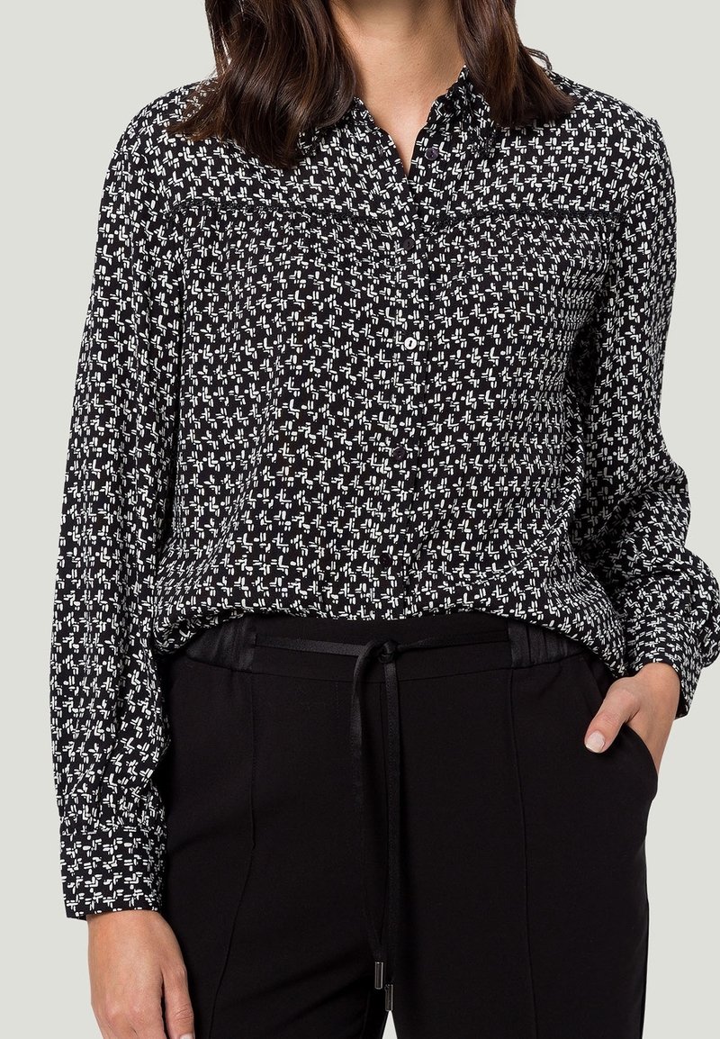 Chemise noire et blanche à boutons avec un motif géométrique, manches longues, et une poche poitrine, associée à un pantalon noir avec un cordon de serrage.