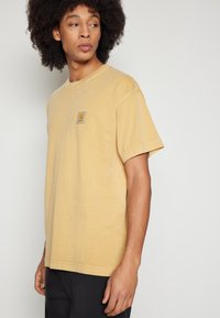 Camiseta de algodón amarilla con mangas cortas, que presenta un pequeño parche con logo en el pecho, de color sólido y un corte relajado. Pantalones negros.