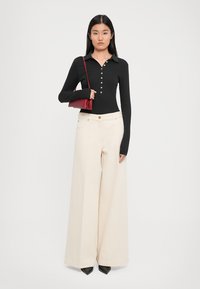 Polo de lungime lungă, de culoare neagră, cu nasturi, asortat cu pantaloni largi de culoare crem. Modelul ține în mână o geantă mică roșie. Țesătură cu un finisaj texturat.