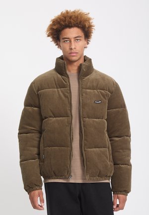 Jeune homme aux cheveux bouclés portant une doudoune en velours côtelé marron sur une chemise beige devant un fond blanc uni.