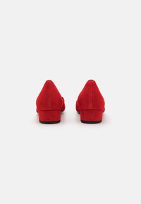 Chaussures à talon bloc en daim rouge avec un bout pointu, présentant une texture lisse et un design minimaliste. Coutures visibles à l'arrière.