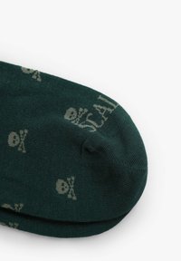 Mørkegrønne strømper med et gentaget mønster af lys beige dødningehoveder og korslagte knogler. Stoffet ser blødt ud med en glat tekstur.