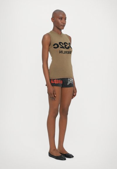 Top senza maniche verde oliva con testo nero "3320 BERLINO" e pantaloni shorts neri con design grafici rossi. Scarpe nere flat completano l'outfit.