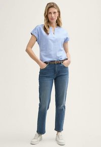 Šviesiai mėlyna trumpomis rankovėmis palaidinė su V formos iškirpte, derinama su tamsiai mėlynais džinsais ir baltais sportbačiais. Diržas su paprasta sagtimi pabrėžia liemenį.