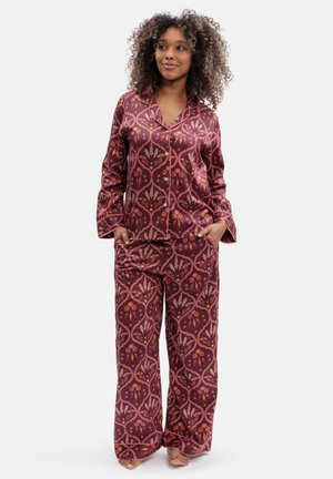 Woman with curly hair wearing matching long-sleeve buttoned top and wide-leg pants with a red and orange patterned design, standing barefoot.