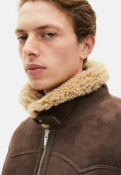 Chaqueta de ante marrón con cuello de shearling en tono beige, que presenta un cierre de cremallera plateado y un distintivo detalle en las costuras. Primer plano del cuello y la cara.