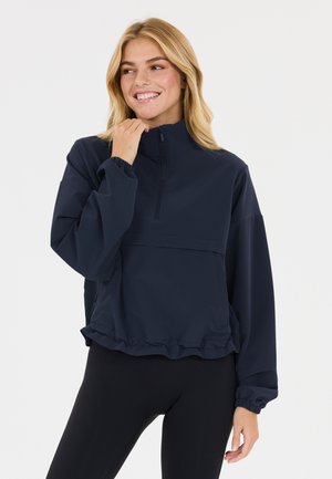 Lyshåret kvinde smilende, iført en marineblå pullover med halv lynlås og elastiske manchetter samt sorte leggings, poserer mod en ensfarvet hvid baggrund.