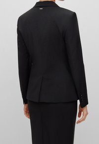 Musta räätälöity takki, jossa on muotoiltu malli, sileä pinta, takana yksittäinen halkio ja neljän napin yksityiskohdat hihoissa. Logo kauluksessa.