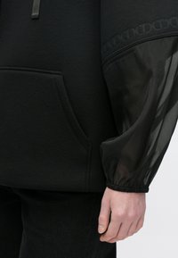 Črna jopa s flisastim telesom in prozornimi, zategnjenimi rokavi z dekorativnim trakom. Vključuje žep spredaj. Minimalističen dizajn.