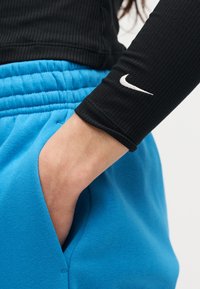 Χέρι ατόμου στην τσέπη μπλε φόρμας, φοράει μαύρο ριμπ μανίκι με λευκό λογότυπο Nike στον καρπό.