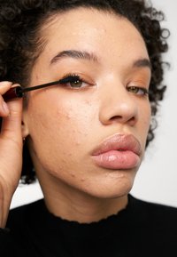 Du mascara noir est appliqué sur un cil recourbé. Le sujet a des cheveux courts et bouclés, un teint moyen et porte un haut noir avec un décolleté festonné.