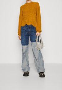 Person in einem senfgelben Kabelstrickpullover, zweifarbigen, weiten Jeans, schwarzen klobigen Schuhen und mit einer kleinen silbernen, strukturierten Handtasche.