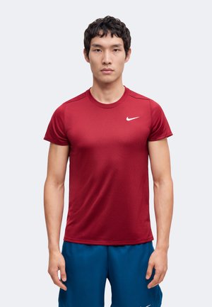 Giovane uomo in piedi di fronte, indossa una maglia rossa a maniche corte Nike e pantaloncini blu su uno sfondo chiaro uniforme.