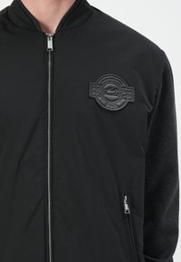 Black bomber jacket with a textured black patch, zip closure, and side pockets. Features a smooth fabric with contrasting knit sleeves.