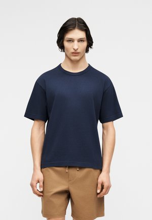 Młoda osoba o średniej długości ciemnych włosach, ubrana w granatowy t-shirt z krótkim rękawem i jasnobrązowe szorty, na jednolitym tle.