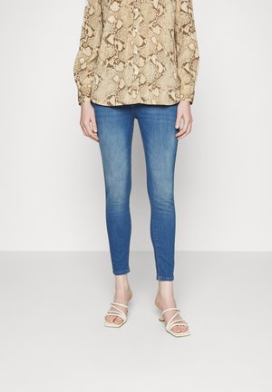 Hellbraun gemusterte Bluse mit langen Ärmeln, kombiniert mit blauen Skinny-Jeans. Weiße, geschnürte Absatzsandalen vervollständigen das Outfit.