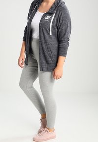 Šedá mikina Nike s bílým logem a šňůrkami, v kombinaci se světle šedými legínami a světle růžovými teniskami. Hladká tkanina, volný střih.