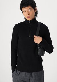 Pull à col roulé noir côtelé avec une fermeture éclair demi-zip. Le modèle tient un petit sac noir. Le tissu semble tricoté et texturé.